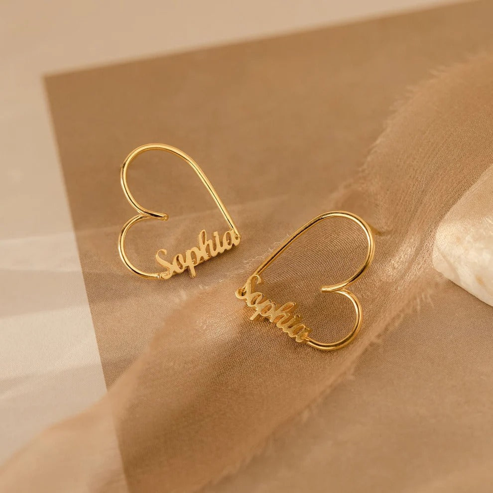 Gold heart-shaped earrings with 'Sophie' inscription on a beige fabric background