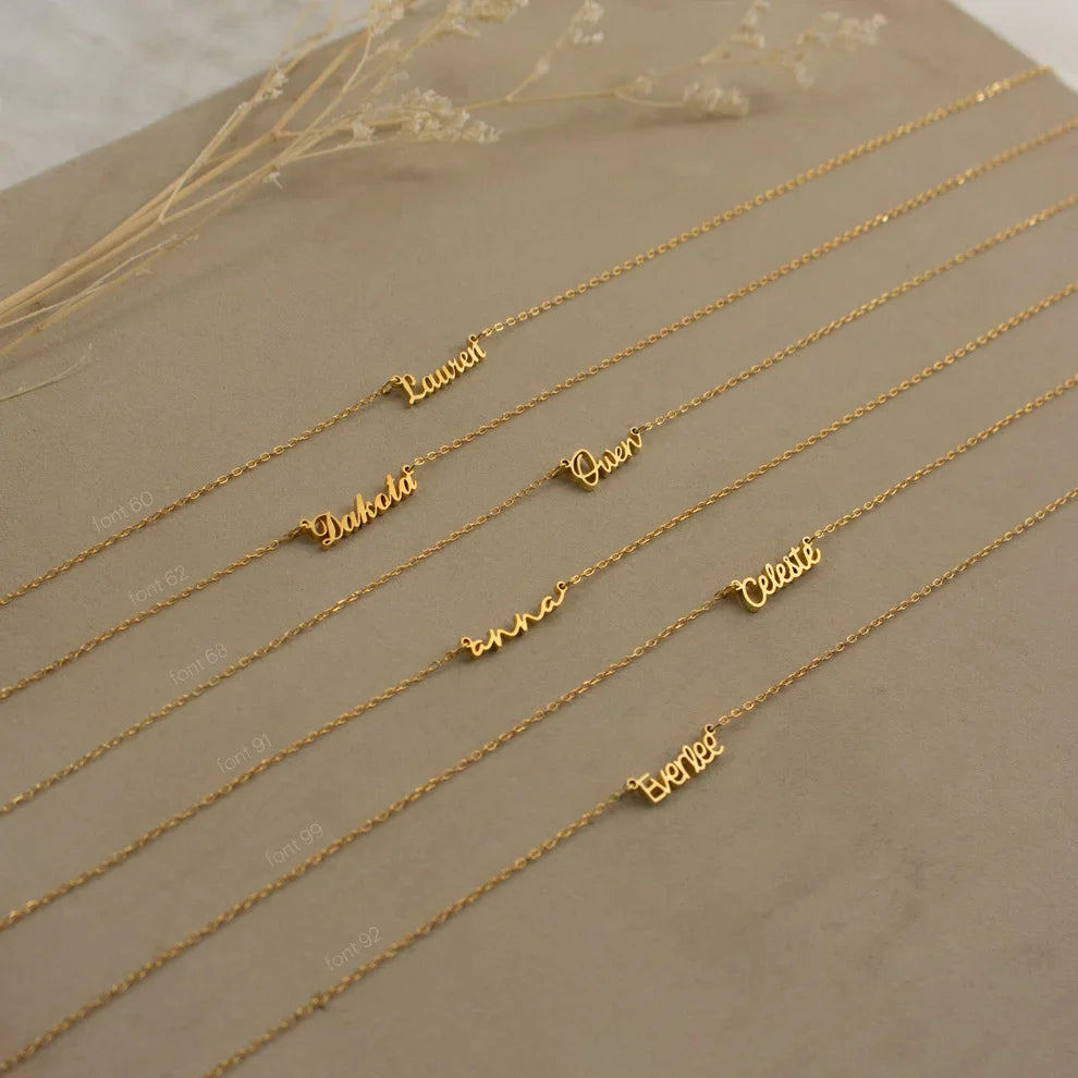 Gold name necklaces on a beige surface with dried flowers.
